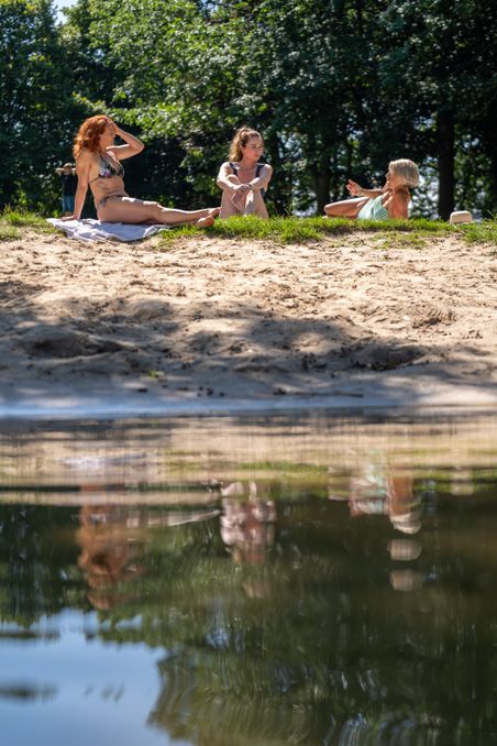 Drei Frauen entspannen sich am sandigen Ufer eines Sees, während sie sich unterhalten. Ihre Spiegelbilder sind im ruhigen Wasser sichtbar.