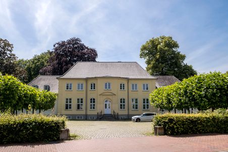 Das Caspar-von-Saldern-Haus in Neumünster ist ein gelbes, historisches Gebäude mit symmetrischer Fassade, umgeben von Bäumen und einem gepflasterten Vorplatz.