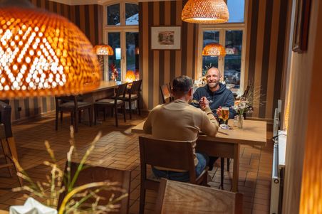 Zwei Männer sitzen im gemütlichen Landgasthof Unter den Linden und unterhalten sich bei Getränken. Die warme Beleuchtung schafft eine einladende Atmosphäre.