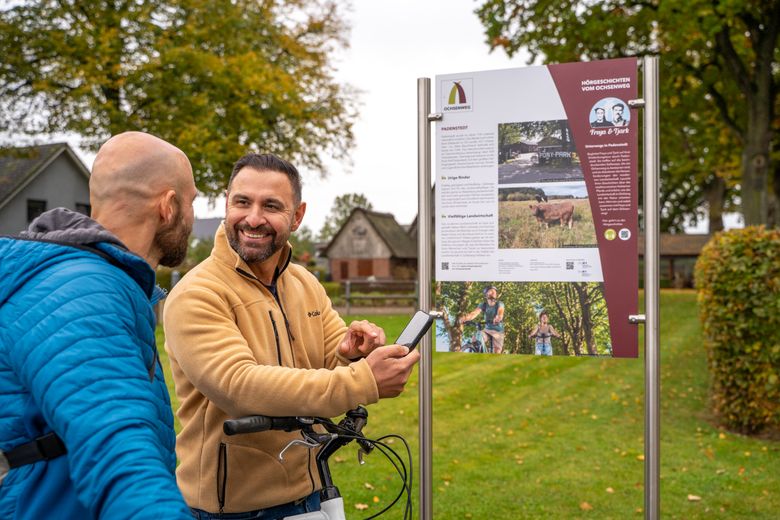 Zwei Männer betrachten eine Informationstafel am Ochsenweg in Padenstedt. Einer hält ein Smartphone, beide wirken interessiert und fröhlich.