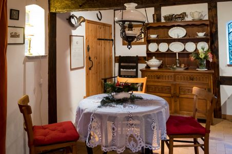 Gemütliche Stube mit Holzmöbeln, Spitzendecke auf dem Tisch und dekorativem Geschirr im Schrank. Einladende Atmosphäre mit traditionellem Charme.