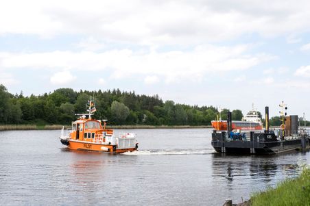 Ein orangefarbenes Lotsenboot auf einem Fluss, begleitet von einem größeren Schiff im Hintergrund. Die Umgebung ist von Bäumen und Gras gesäumt.