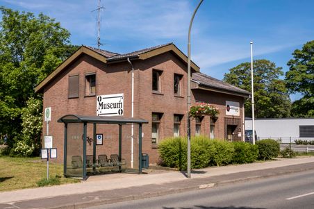 Backsteingebäude mit dem Schild 'Museum' und einer Bushaltestelle davor. Das Gebäude ist von Bäumen und Sträuchern umgeben.