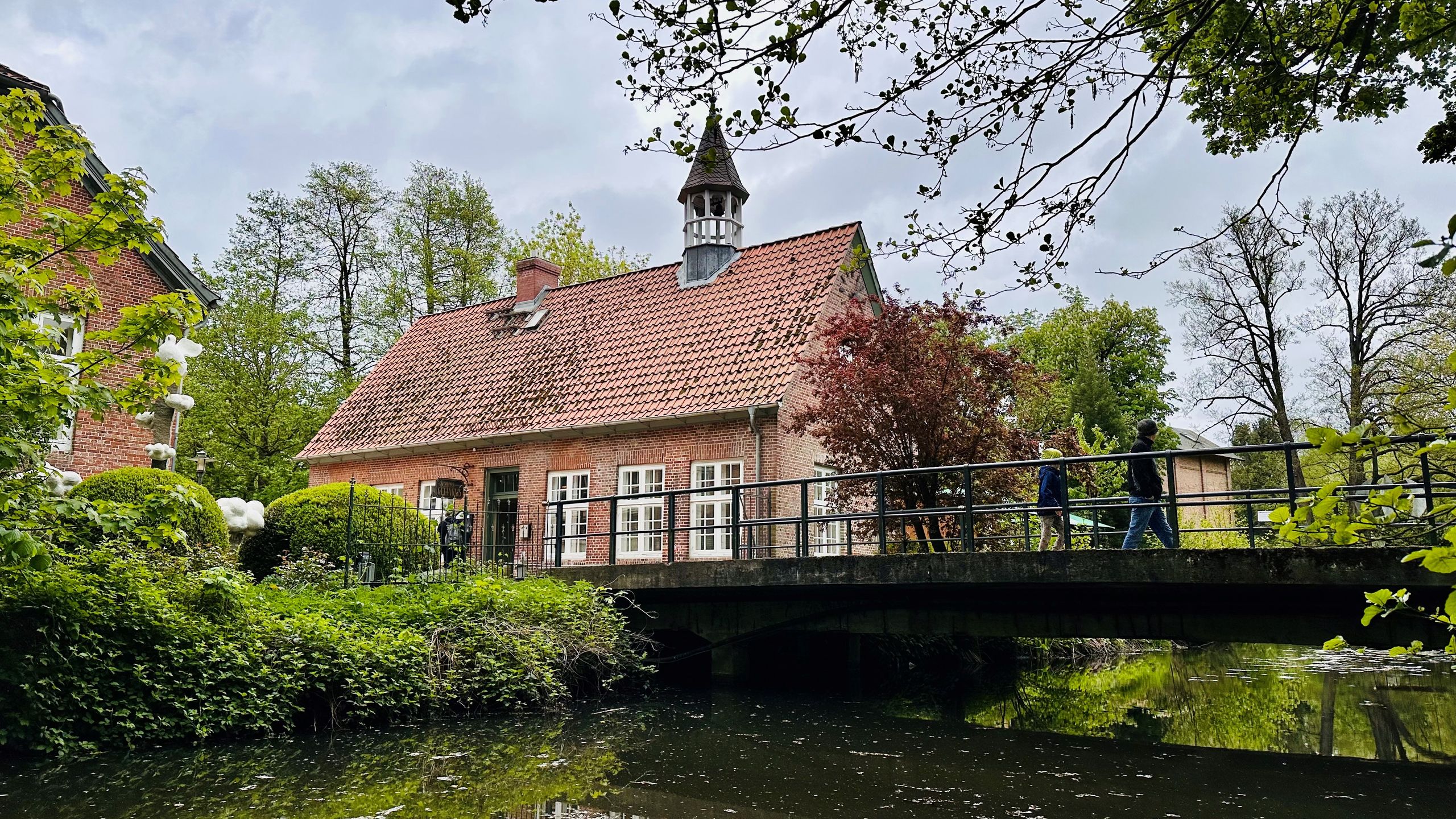 Ein malerisches Backsteingebäude mit Turm, umgeben von grüner Natur und einem Fluss. Eine Brücke führt über das Wasser, während Menschen darauf spazieren.