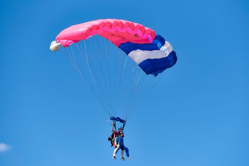 Zwei Personen machen einen Tandem-Fallschirmsprung unter einem pink-blauen Fallschirm vor klarem, blauem Himmel.