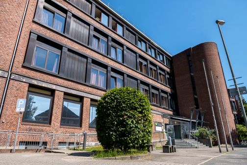 Ein modernes Backsteingebäude mit großen Fenstern und einem runden Treppenhaus in Neumünster, umgeben von einem gepflasterten Vorplatz und einem Baum.