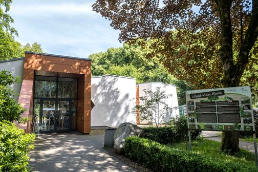 Eingang des Tierparks Neumünster mit moderner Architektur und einem Informationsschild. Umgeben von Bäumen und grüner Vegetation.