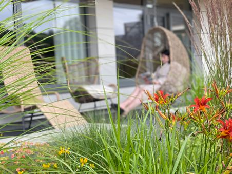 Rote Blumen und grünes Gras im Vordergrund, während im Hintergrund eine Person in einem Hängesessel auf einer Terrasse entspannt.