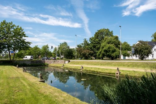 Ein kleines Gewässer mit Statuen am Ufer, umgeben von grünen Bäumen und einer Straße im Hintergrund. Der Himmel ist blau mit einigen Wolken.