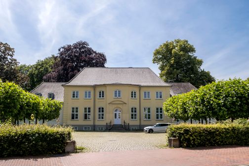 Das Caspar-von-Saldern-Haus in Neumünster ist ein gelbes, historisches Gebäude mit symmetrischer Fassade, umgeben von Bäumen und einem gepflasterten Vorplatz.