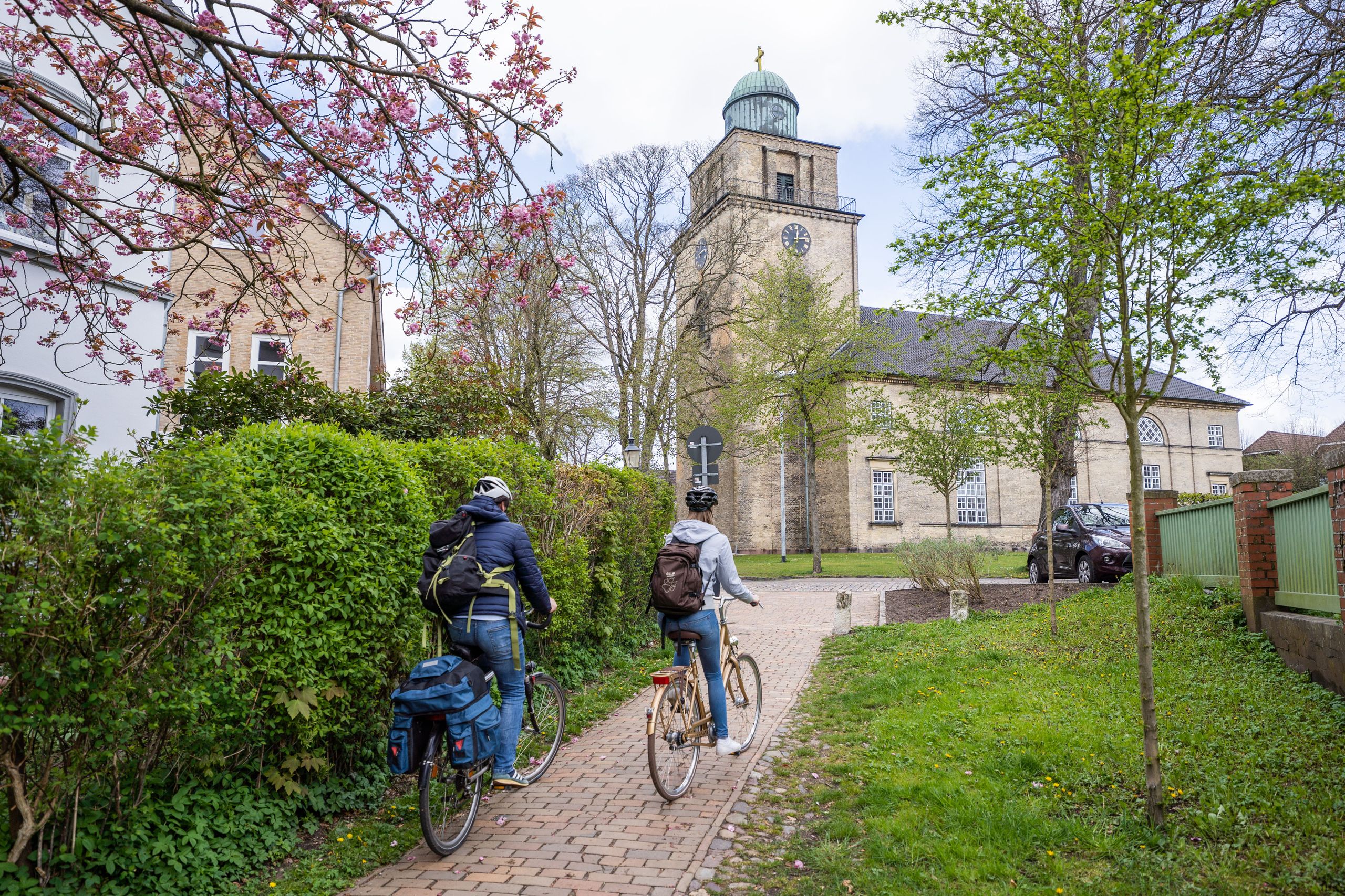 Zwei Radfahrer fahren auf einem gepflasterten Weg zur Vicelinkirche in Neumünster. Blühende Bäume und grüne Büsche säumen den Weg.