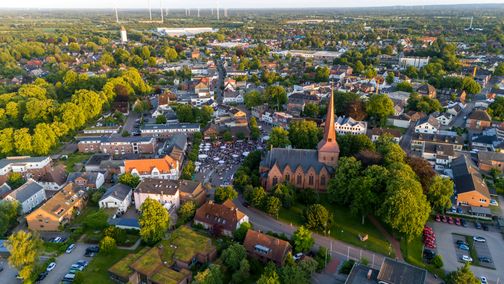 Luftaufnahme von Nortorf zeigt eine Kirche im Zentrum, umgeben von Wohnhäusern und viel Grün. Im Hintergrund sind Windräder zu sehen.
