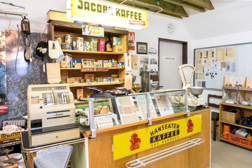 Ein nostalgischer Laden mit alter Registrierkasse, Waage und Regalen voller Vintage-Produkte. Ein 'Jacobs Kaffee'-Schild hängt über dem Tresen.