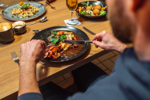 Ein Mann genießt ein Gericht mit Fleisch, Gemüse und Soße. Im Hintergrund sind weitere Teller mit Speisen und ein Glas Bier zu sehen.