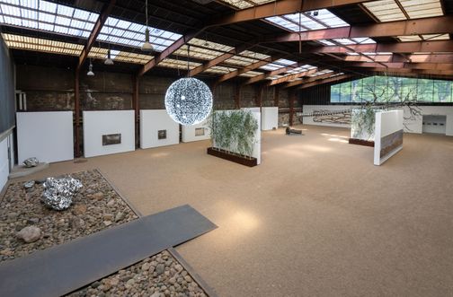 Große Ausstellungshalle mit moderner Kunstinstallation. Eine leuchtende Kugel hängt von der Decke, umgeben von Skulpturen und Pflanzenwänden.