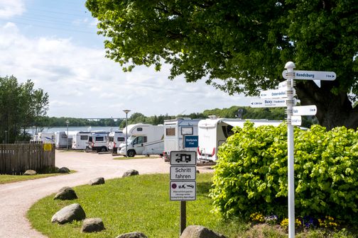 Wohnmobilstellplatz in Schacht-Audorf mit mehreren Wohnmobilen, einem Wegweiser und einem Schild, das zur langsamen Fahrt auffordert.
