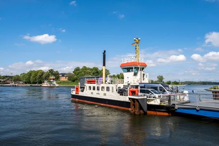 Eine Fähre transportiert Autos über einen Fluss. Im Hintergrund sind Bäume und Gebäude zu sehen. Der Himmel ist blau mit einigen Wolken.