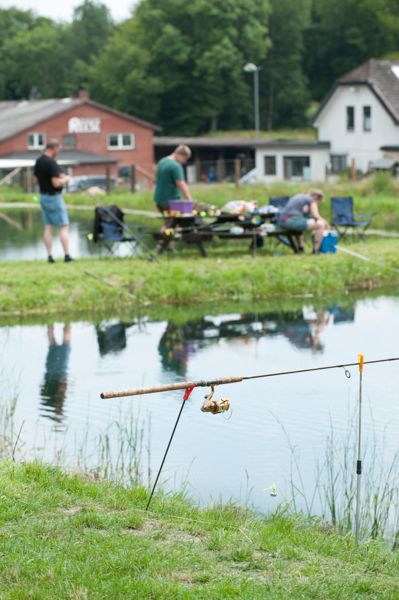 Angelrute am Teichufer, im Hintergrund mehrere Personen beim Beisammensein vor Häusern und Bäumen.