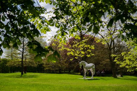 Eine weiße Pferdeskulptur auf einer grünen Wiese, umgeben von Bäumen. Menschen stehen im Hintergrund. Die Szene wirkt friedlich und naturverbunden.