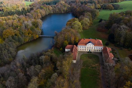 Luftaufnahme von Gut Emkendorf, umgeben von herbstlichen Bäumen und einem See. Das Herrenhaus hat ein rotes Dach und liegt in einer grünen Landschaft.