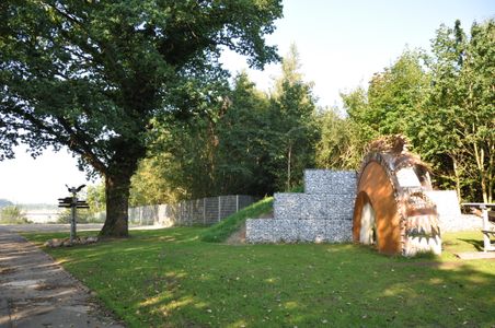 Ein grüner Aussichtspunkt mit einem großen Baum, einem Wegweiser und einer Skulptur aus Metall und Steinen. Im Hintergrund sind Bäume und ein Zaun zu sehen.
