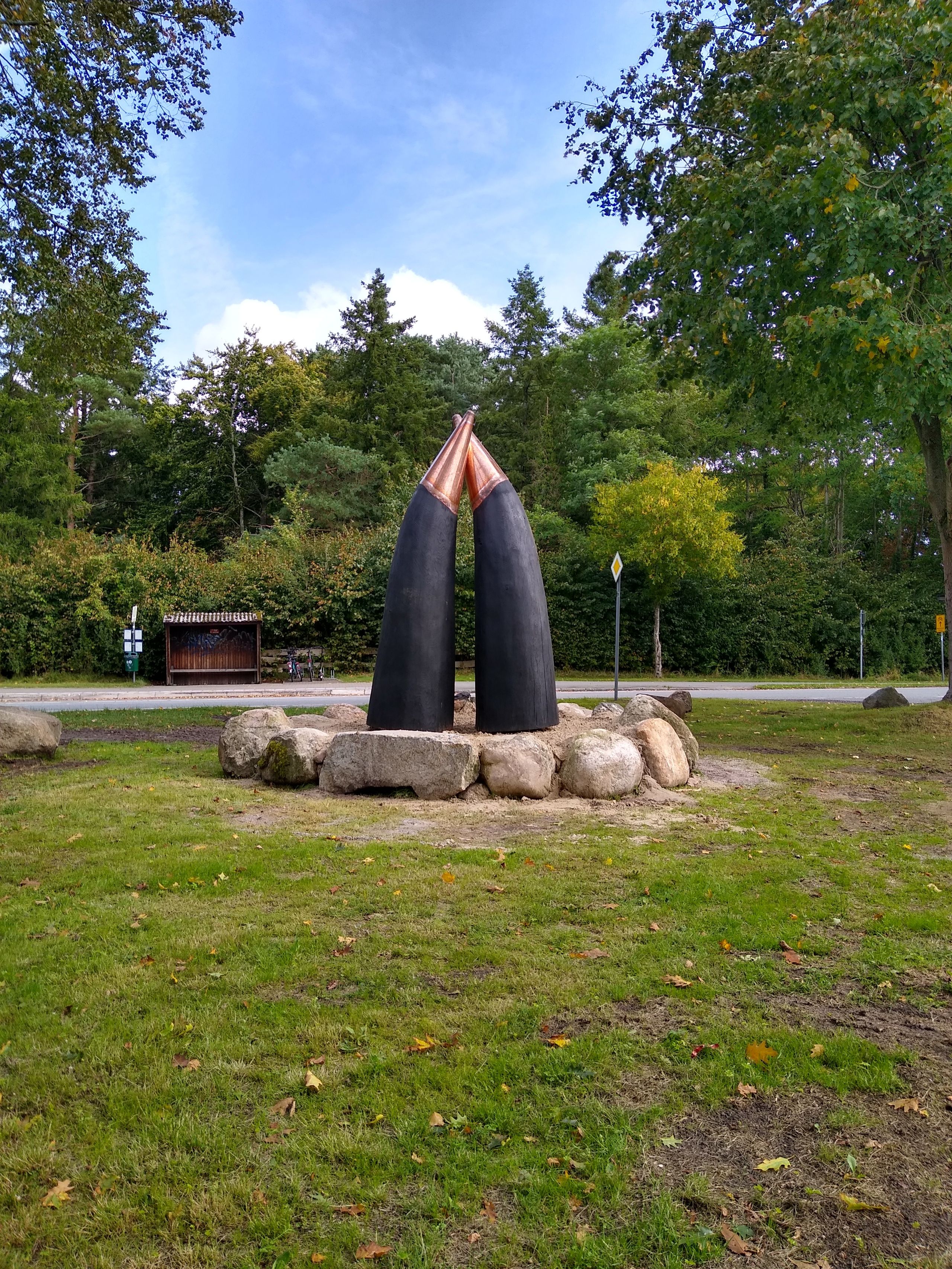 Zwei große, schwarze Hörner stehen auf einem Steinkreis in einem grünen Park. Im Hintergrund sind Bäume und ein blauer Himmel zu sehen.