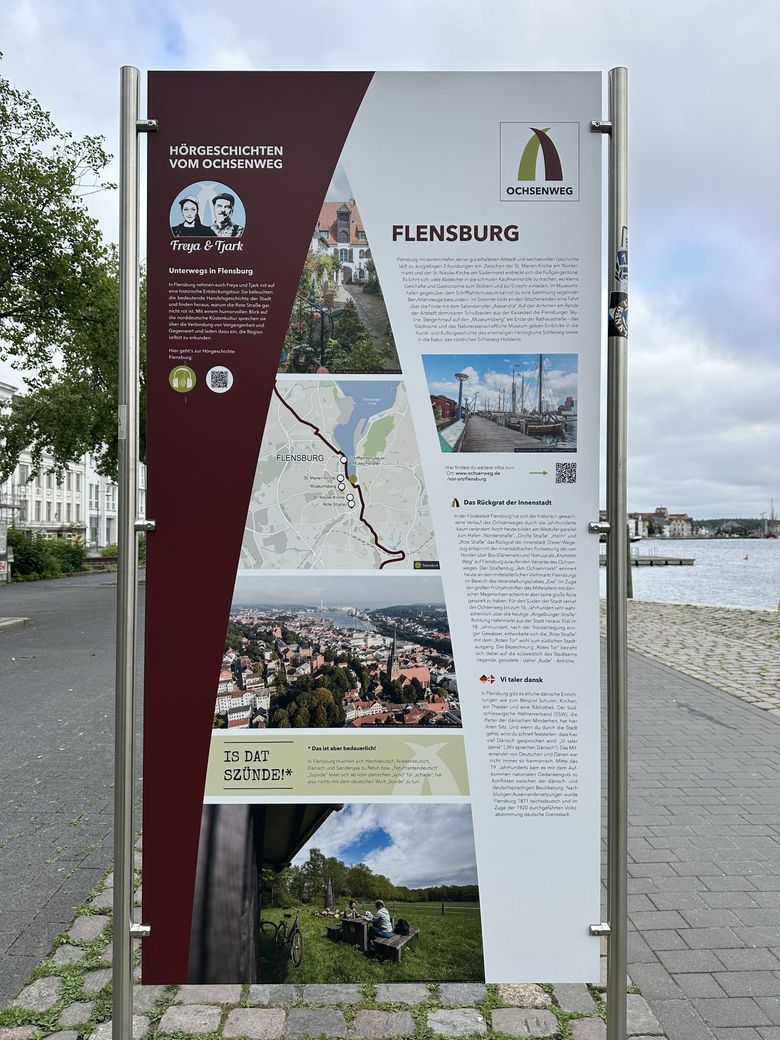 Eine Infotafel in Flensburg beschreibt den Ochsenweg mit Bildern, Texten und einer Karte. Es gibt Hinweise auf Hörgeschichten und lokale Sehenswürdigkeiten.