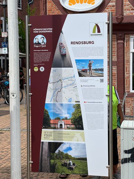 Ein Informationsschild in Rendsburg über den Ochsenweg mit Bildern, Texten und einer Karte. Es steht auf einem gepflasterten Platz vor einem Backsteingebäude.