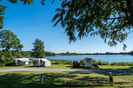 Wohnmobile stehen auf einem grünen Campingplatz am Seeufer in Borgdorf-Seedorf. Der Himmel ist klar und blau, umgeben von Bäumen.