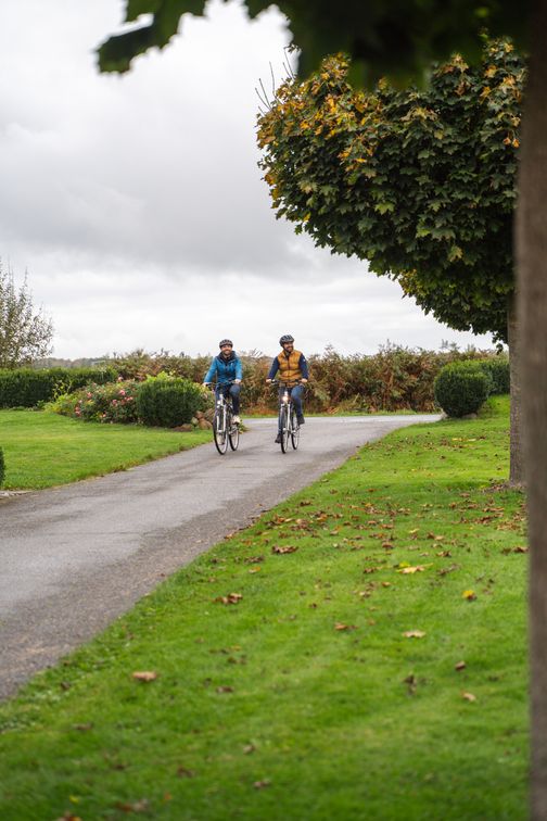Zwei Radfahrer auf einem Weg, umgeben von grüner Landschaft und Bäumen. Der Himmel ist bewölkt, und es ist Herbst.
