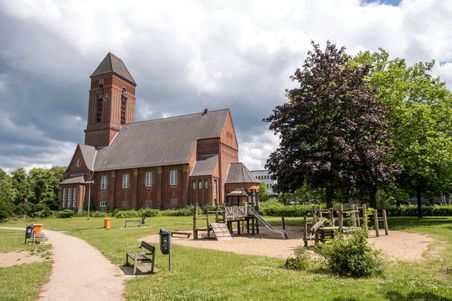 Die Anscharkirche im Anscharpark mit einem Spielplatz im Vordergrund. Der Himmel ist bewölkt, und Bäume umgeben die Szene.