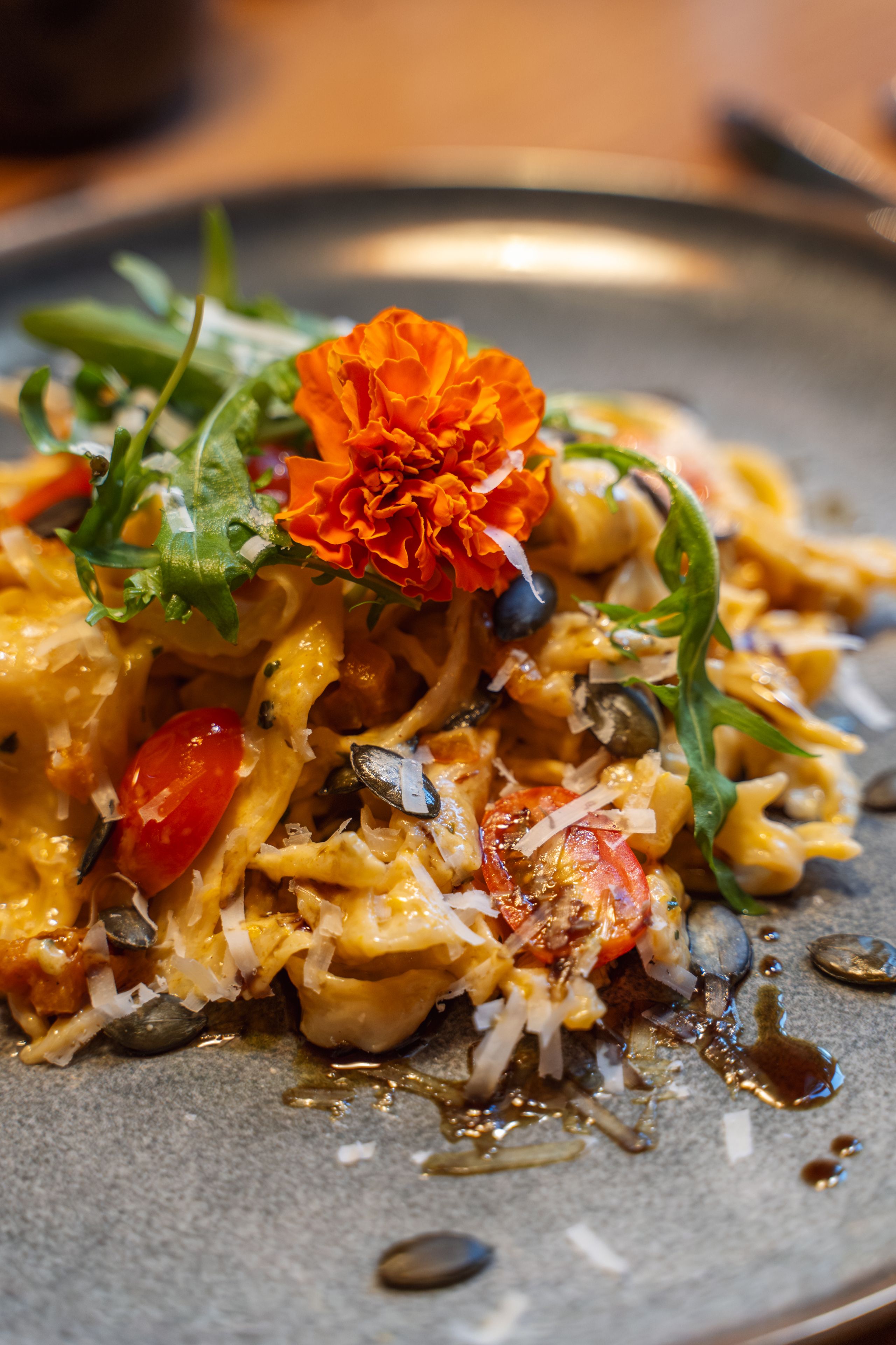 Kreativ angerichtete Pasta mit Blüte, Rucola, Kürbiskernen und Kirschtomaten auf einem grauen Teller.
