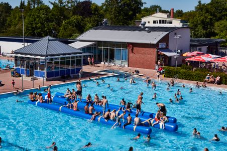 Das Roland Oase Freibad mit vielen Badegästen, aufblasbarem Trimaran und Bistrogebäude an einem sonnigen Tag.