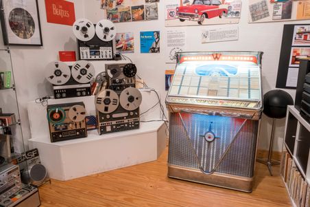 Ein Raum mit mehreren Tonbandgeräten und einer Wurlitzer-Jukebox. An den Wänden hängen Poster und Schallplatten-Cover.