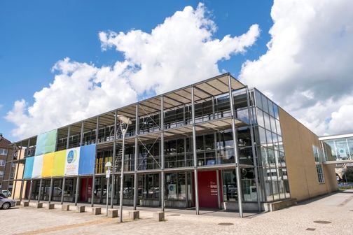 Das Museum Tuch & Technik in Neumünster zeigt eine moderne Glasfassade mit bunten Bannern. Der Himmel ist blau mit weißen Wolken.