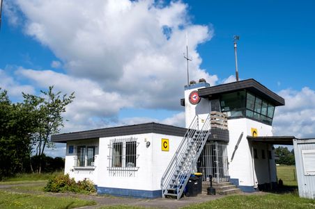 Der Kontrollturm des Flugplatzes Neumünster steht unter blauem Himmel mit weißen Wolken. Ein gelbes Schild mit der Aufschrift 'C' ist sichtbar.