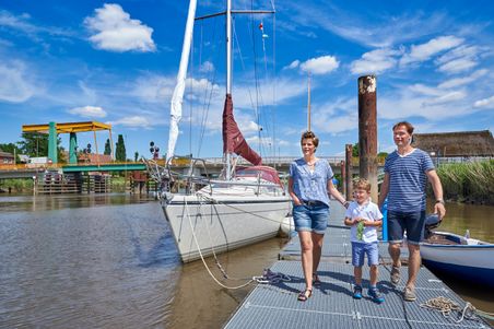 Eine Familie spaziert auf einem Steg neben einem Segelboot im Hafen von Heiligenstedten. Im Hintergrund ist die Klappbrücke zu sehen.