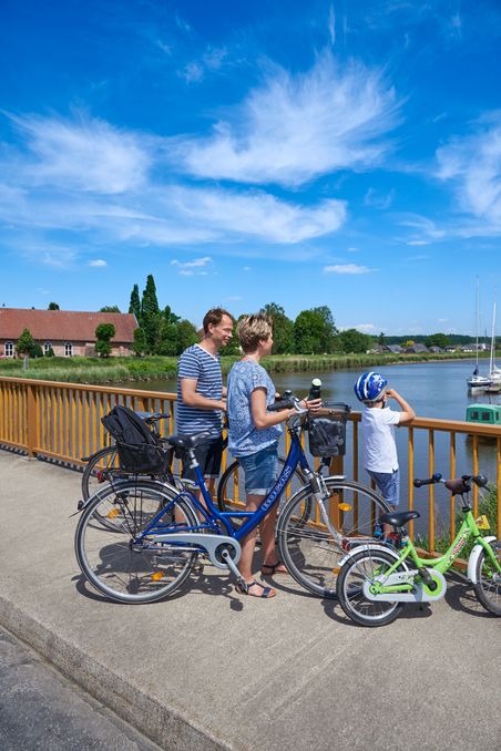 Eine Familie mit Fahrrädern genießt den Ausblick von einer Brücke über einen Fluss. Der Himmel ist klar und blau.