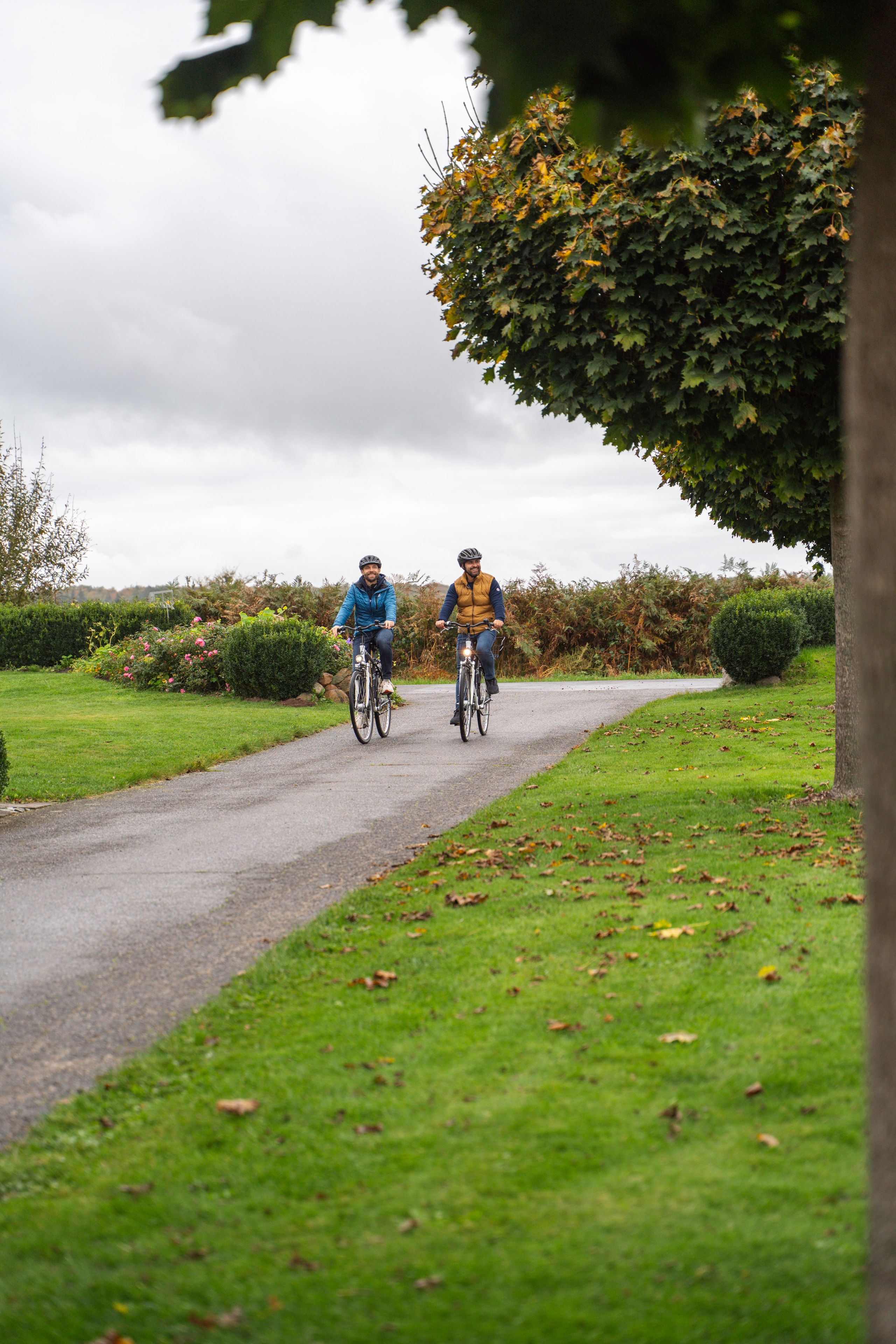 Zwei Radfahrer auf einem Weg, umgeben von grüner Landschaft und Bäumen. Der Himmel ist bewölkt, und es ist Herbst.