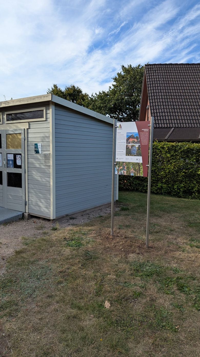 Ein kleines graues Gebäude mit einem Informationsschild zum Ochsenweg daneben. Der Himmel ist blau mit einigen Wolken. Im Hintergrund sind Bäume und ein Hausdach zu sehen.