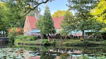 Ein malerisches Backsteingebäude mit rotem Dach am Ufer eines Teiches, umgeben von üppigem Grün und Seerosen im Wasser.