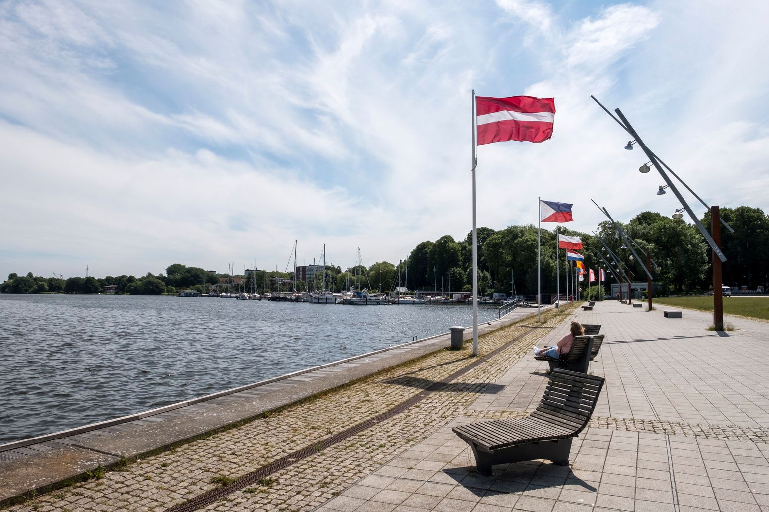 Promenade am Obereiderhafen in Rendsburg mit Flaggen und Bänken. Im Hintergrund sind Boote und Bäume zu sehen, während eine Person auf einer Bank sitzt.
