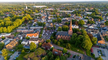 Luftaufnahme von Nortorf zeigt eine Kirche im Zentrum, umgeben von Wohnhäusern und viel Grün. Im Hintergrund sind Windräder zu sehen.