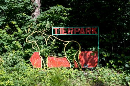 Ein Schild mit der Aufschrift 'Tierpark' und einem Hirschmotiv steht im Grünen, umgeben von dichtem Wald.