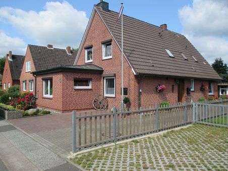 Außenansicht eines roten Backsteinhauses mit Satteldach, Vorgarten und Holzzaun an einer Wohnstraße.