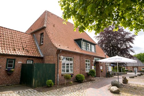 Die Gaststätte Schülper Kroog zeigt ein traditionelles Backsteinhaus mit rotem Ziegeldach. Ein Gerolsteiner Sonnenschirm steht im Außenbereich.