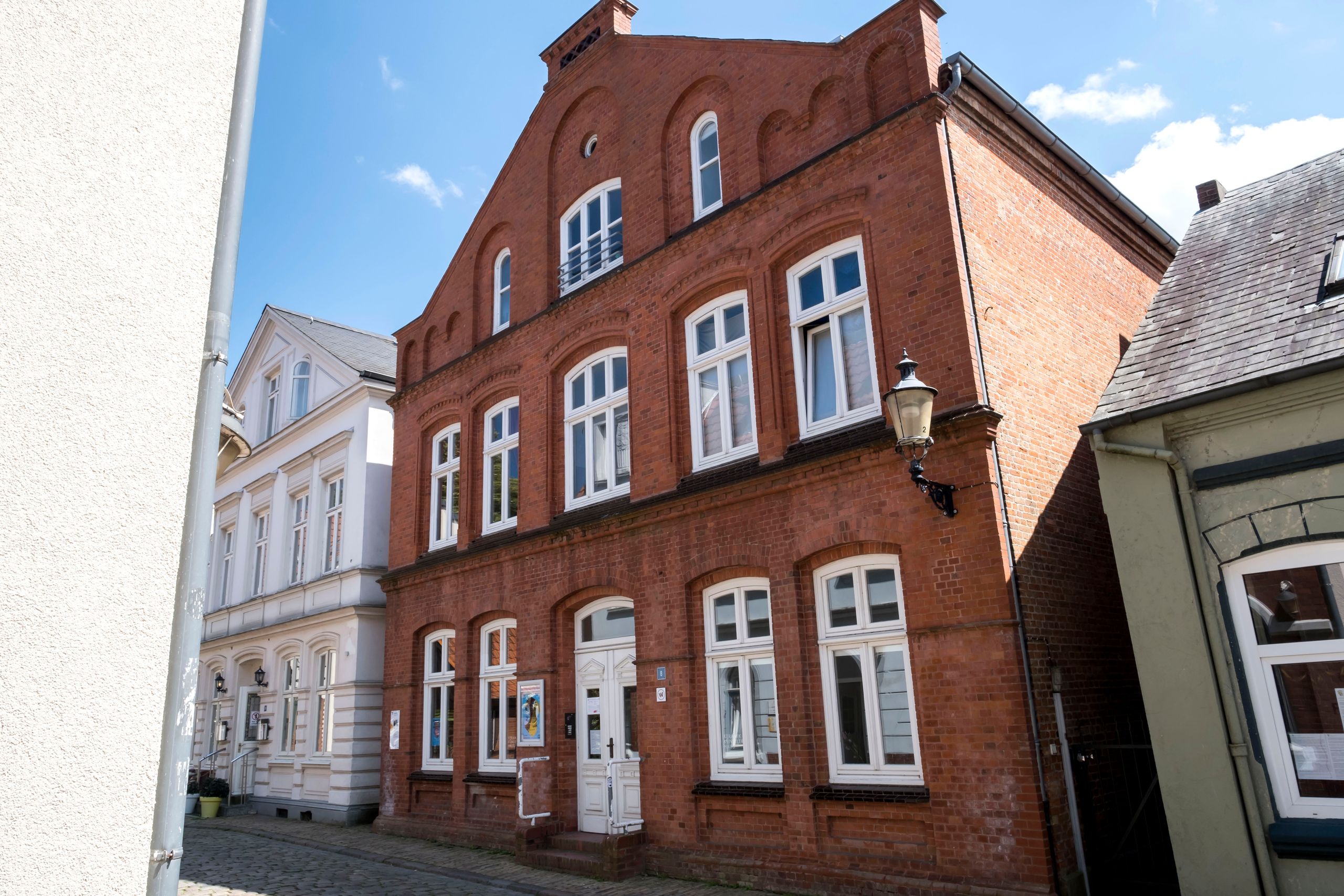 Ein rotes Backsteingebäude mit weißen Fenstern und einer Laterne an der Fassade. Daneben steht ein weißes Gebäude. Der Himmel ist blau mit wenigen Wolken.