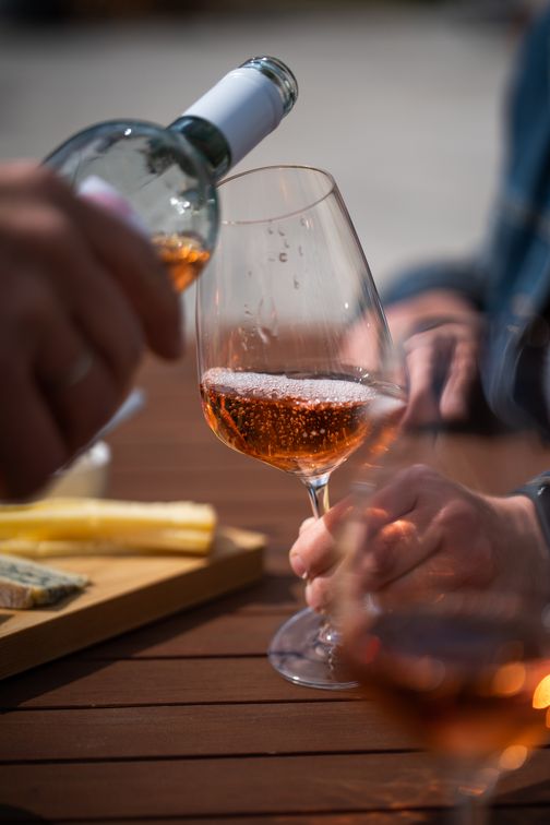 Ein Glas Roséwein wird auf einem Holztisch eingeschenkt. Im Hintergrund sind Hände und ein Käsebrett zu sehen.