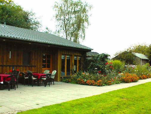 Außenansicht eines Holzgebäudes mit Terrasse, Tischen und Stühlen, Blumenbeet und Rasen.