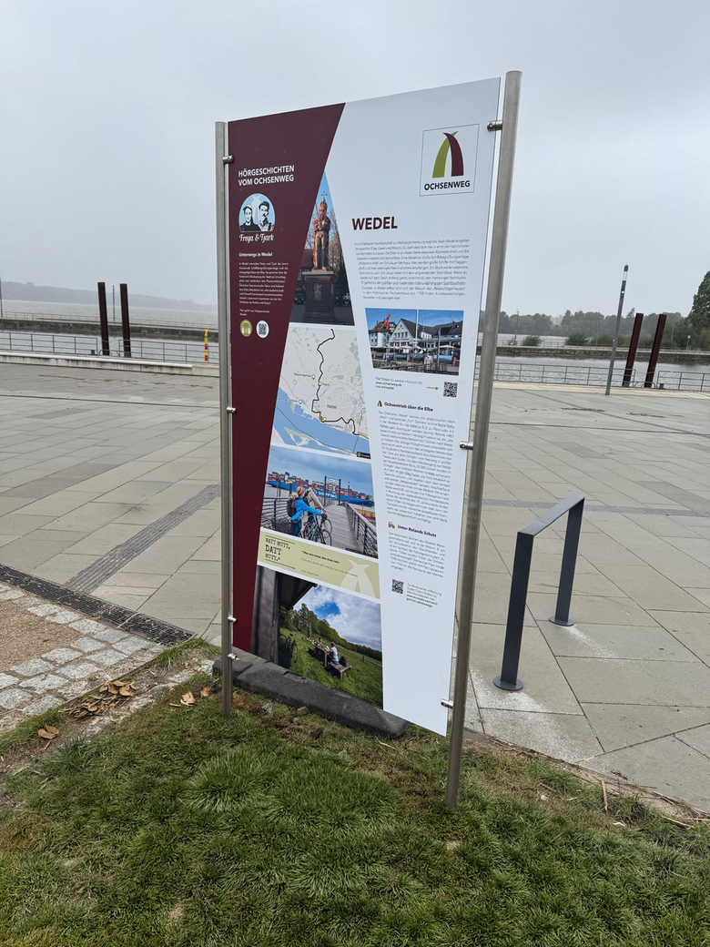Ein Informationsschild in Wedel steht auf einer gepflasterten Fläche mit Blick auf die Elbe. Es beschreibt den Ochsenweg und Sehenswürdigkeiten der Region.